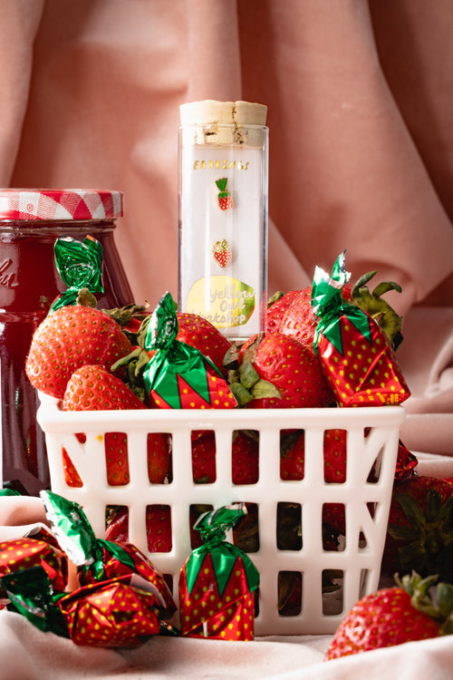 Yellow Owl Workshop strawberry candy earrings in glass vial packaging, styled in a basket of fresh strawberries and vintage strawberry-wrapped hard candies. Nostalgic food-inspired jewelry celebrating the classic strawberry sweets found in grandma's candy dish.