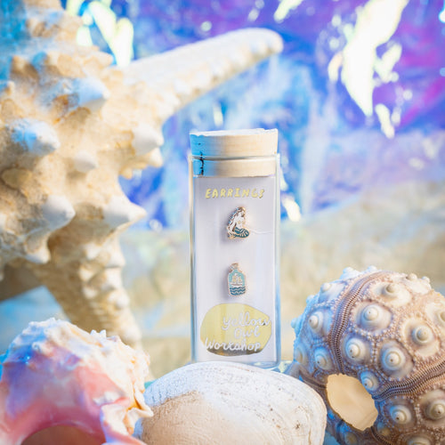 Yellow Owl Workshop mermaid and message in a bottle stud earrings in glass vial packaging, styled among seashells, sea urchins, and pearls against an iridescent blue and purple background. Ocean-inspired mismatched earrings with a coastal, under-the-sea aesthetic.