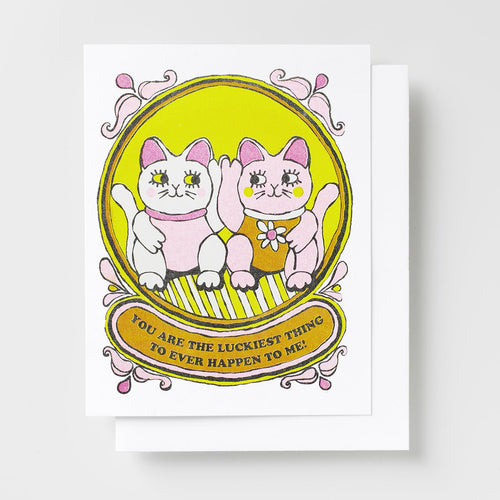 Risograph greeting card with two illustrated lucky cat characters sitting together in an ornate golden oval frame, text reads You Are the Luckiest Thing to Ever Happen to Me on a gold banner