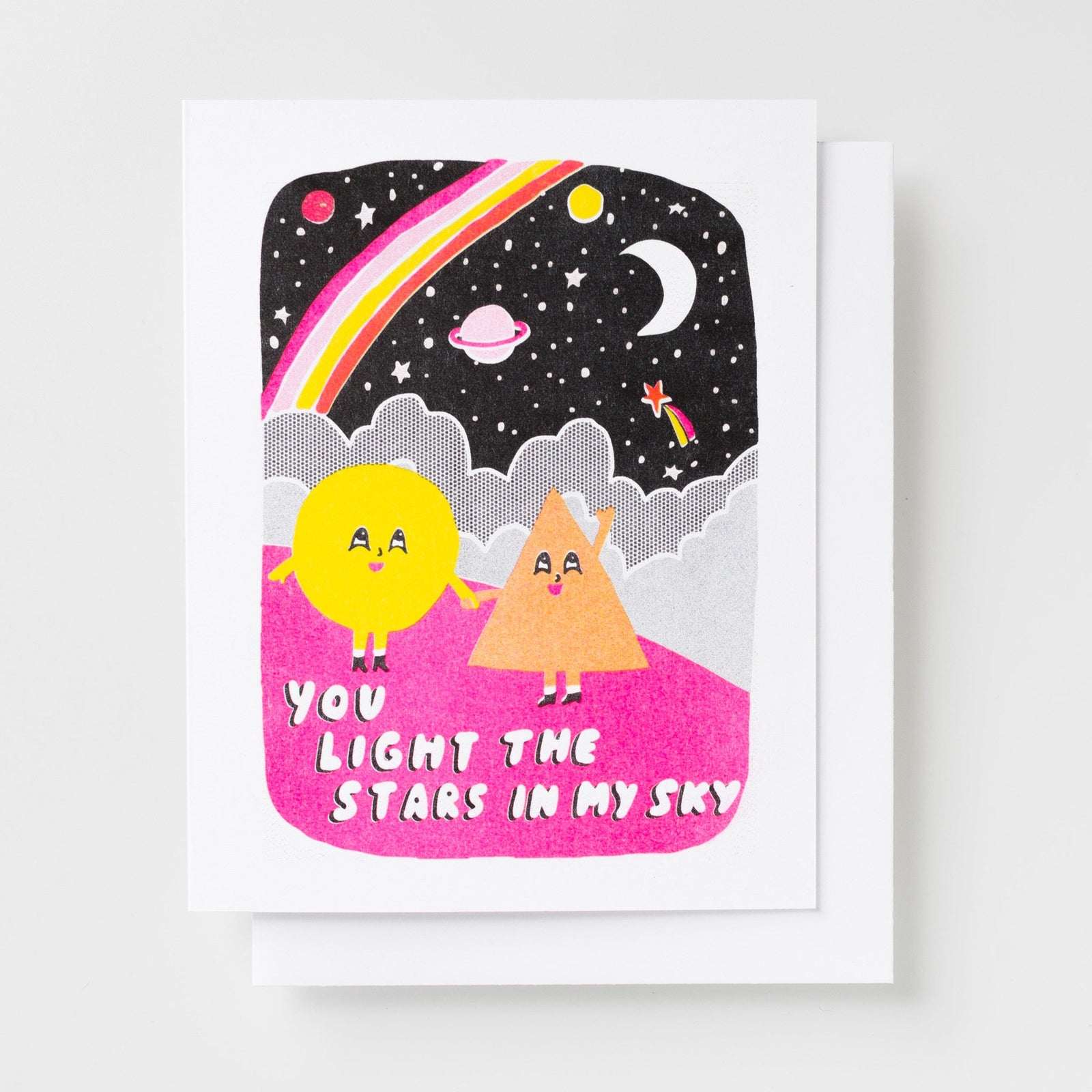 Risograph greeting card with two illustrated characters, a yellow circle and an orange triangle, holding hands on a pink hill beneath a starry night sky with a rainbow, moon, and planets, text reads You Light the Stars in My Sky in white bold lettering